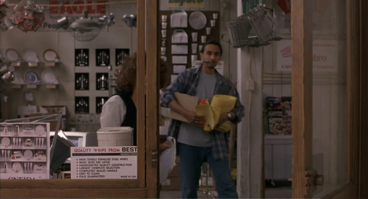 Bogus (1996) movie still: medium — A cluttered wholesale supply shop features shelves of kitchenware and signage on display. A man holding a stack…