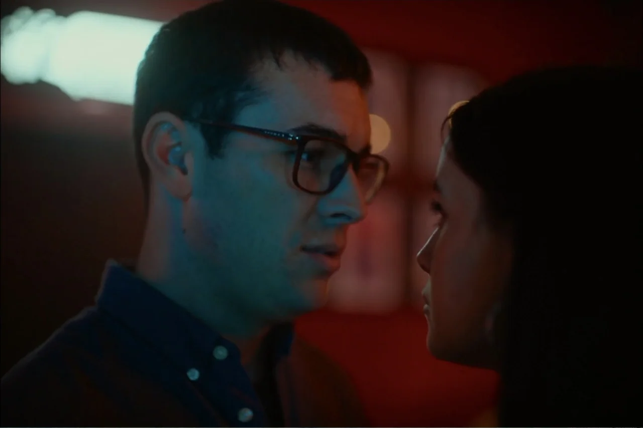 Cross the Line (2020) movie still: closeup — Two individuals stand in a dark interior space with red-toned background elements. A man in glasses and…