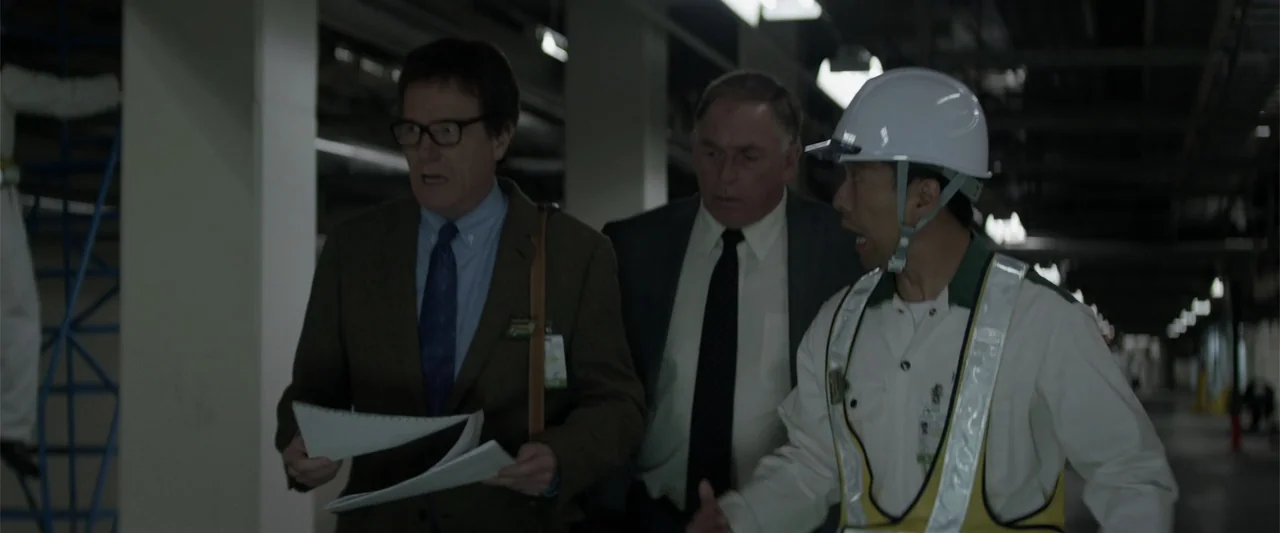Godzilla (2014) movie still: medium — A spacious industrial warehouse interior features white structural columns and metal scaffoldings. Three men…