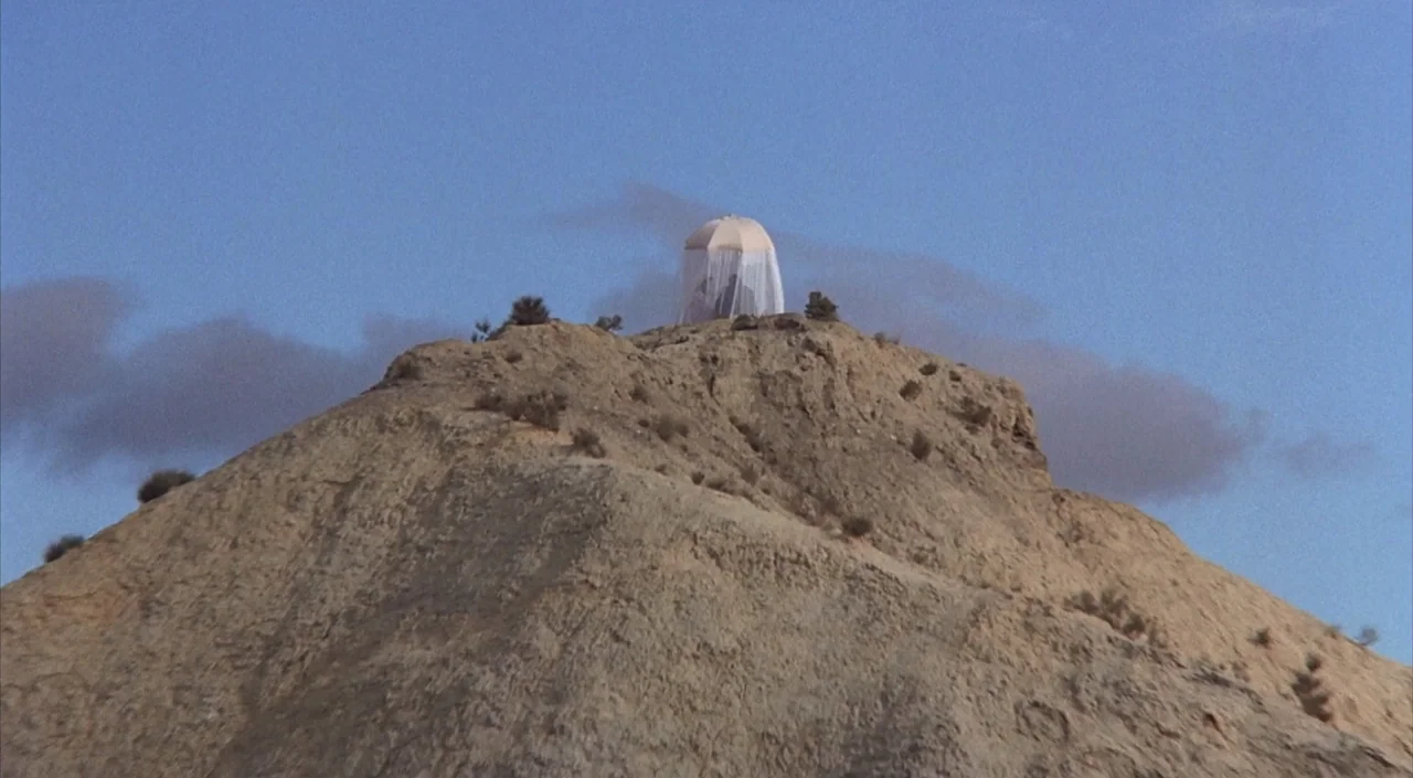 Dr. Phibes Rises Again (1972) movie still: wide — A dry desert hill features a white umbrella with mesh drapes atop the peak. The hill has sparse…