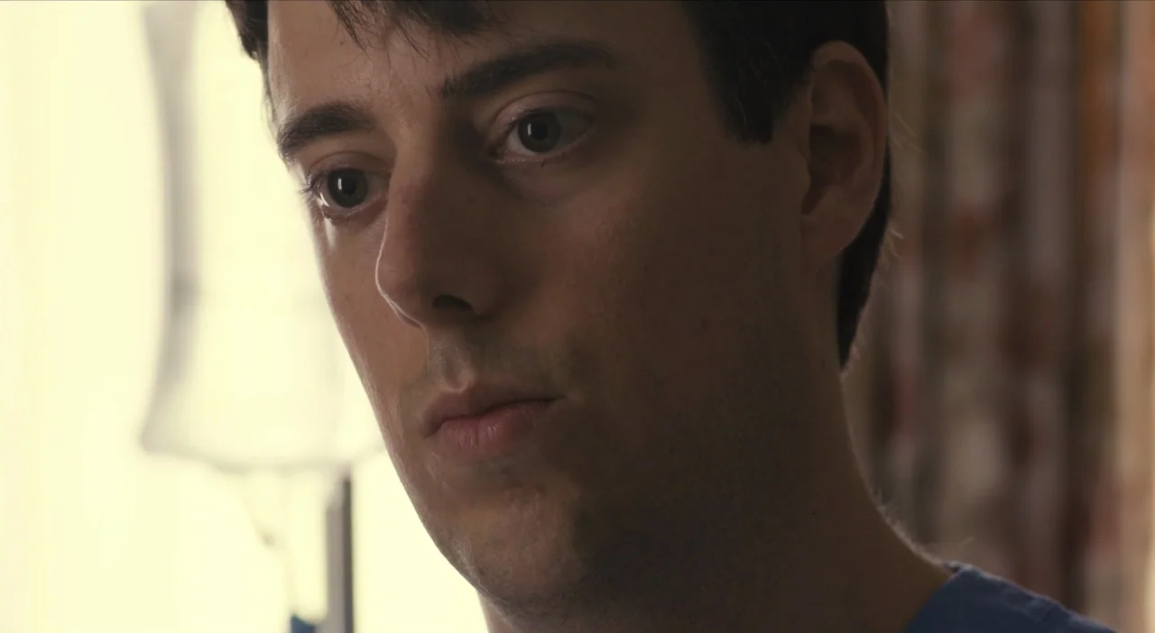 The Comedy (2012) movie still: closeup — An interior space with a window out of focus in the bright background. A young man stares past the camera…