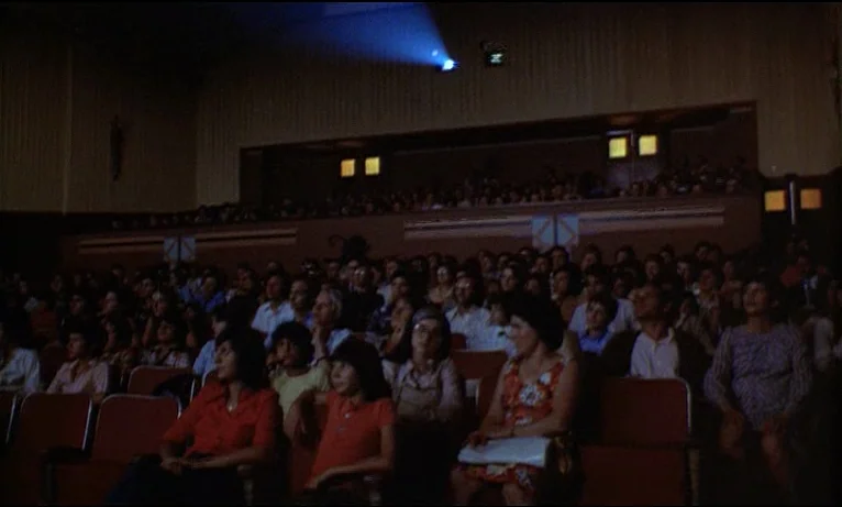 Small Change (1976) movie still: wide — A crowded auditorium contains rows of orange-colored seats occupied by an audience. A bright blue beam of…