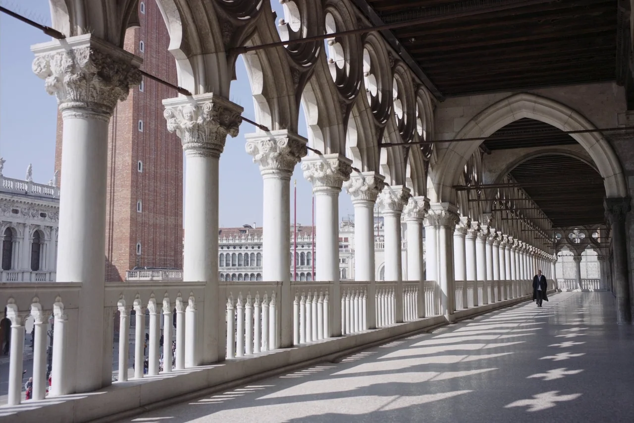 Anselm (2023) movie still: wide — A long marble loggia with repeating white arches overlooking a historic city square. A single figure in dark…