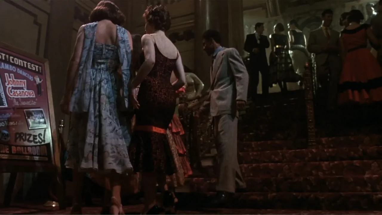 The Mambo Kings (1992) movie still: medium — A dimly lit grand staircase features patrons in 1950s attire moving toward the entrance. A contest…