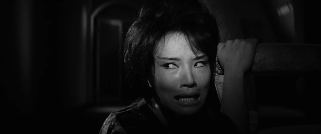 Branded To Kill (1967) movie still: closeup — A dark interior with a blurred archway in the background. A woman looks off-frame with her teeth bared…