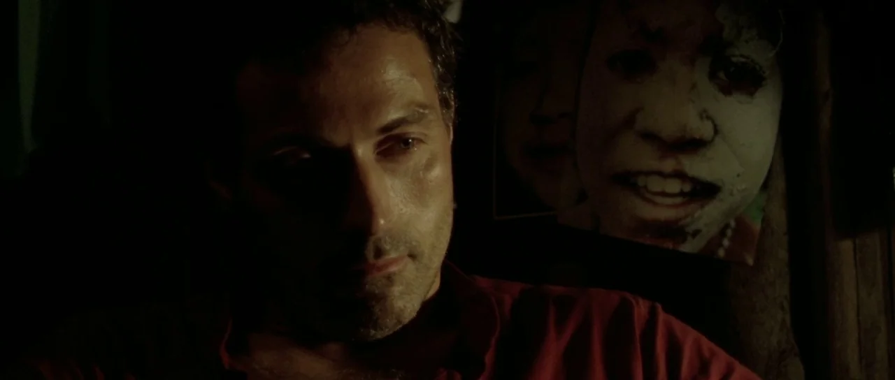 Vinyan (2008) movie still: closeup — A dimly lit room features a man partially illuminated against a dark background. A faded poster of a person…