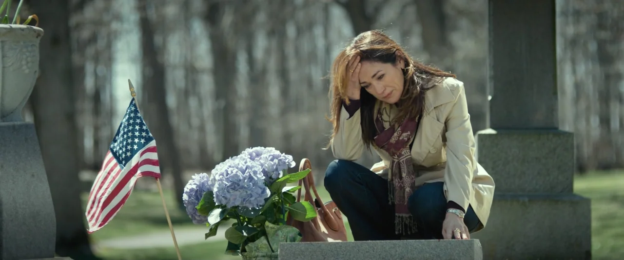 God Bless the Broken Road (2018) movie still: medium — A rural cemetery with gray stone headstones and blurred trees in the background. A woman in a…