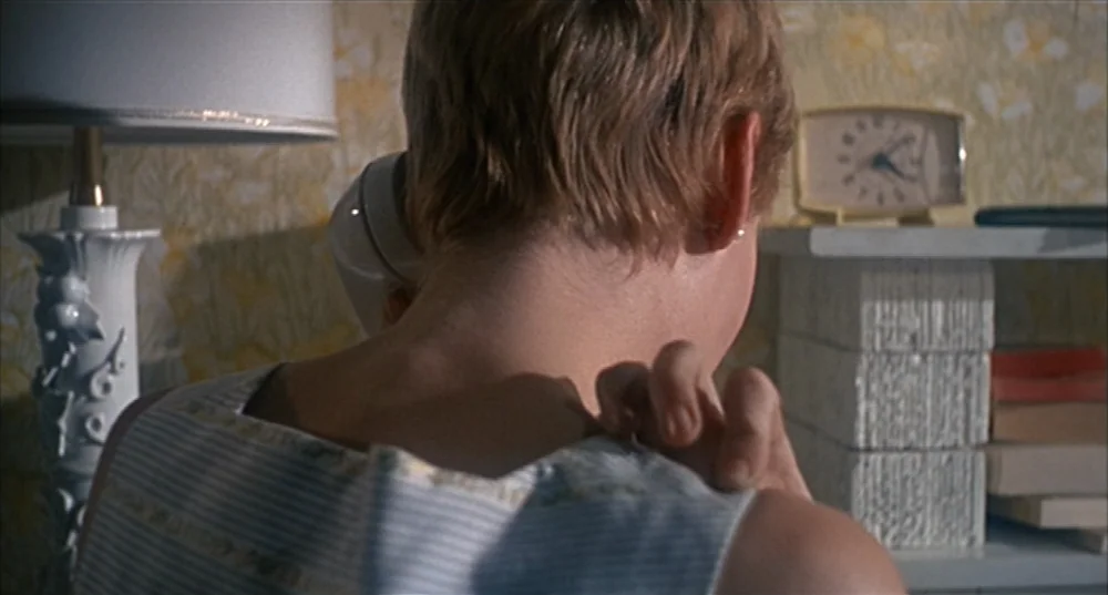 Rosemary’s Baby (1968) movie still: closeup — A 1960s hotel room features yellow floral wallpaper and a small bedside shelf unit. A woman with short…