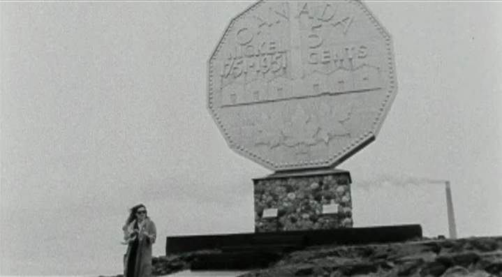 Roadkill (1989) movie still: wide — A massive concrete coin monument stands atop a field on a cloudy day. A woman in a trench coat walks across the…