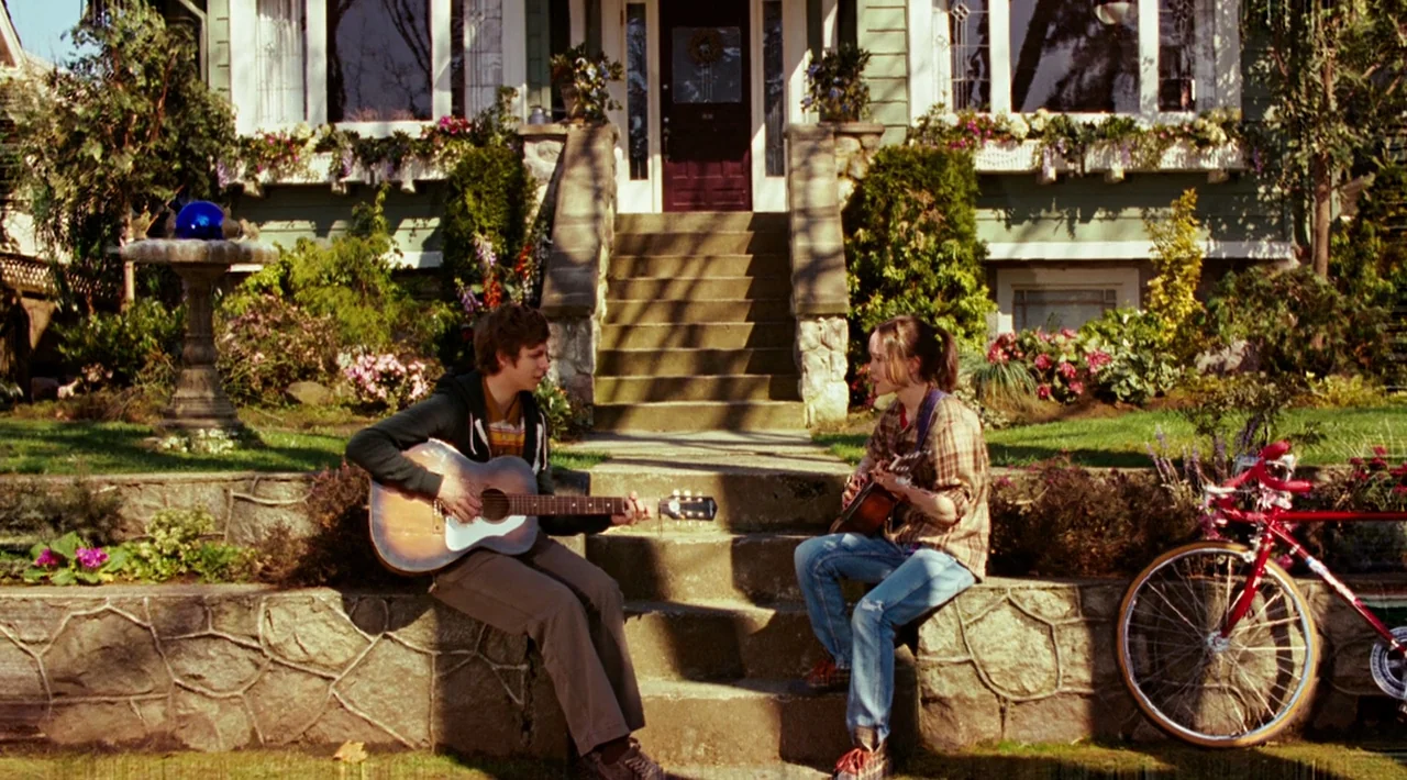 Juno (2007) movie still: fullbody — A house front with stone steps and a landscaped yard is bathed in sunlight. A young man and woman sit on the…
