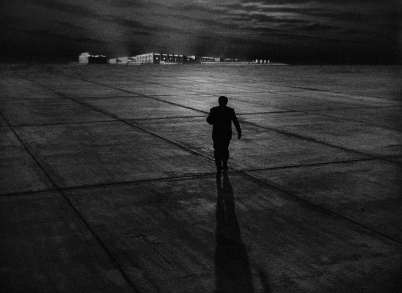 Krakatit (1948) movie still: wide — A vast, empty concrete tarmac stretches toward a lit complex of industrial buildings. A lone man in a suit walks…
