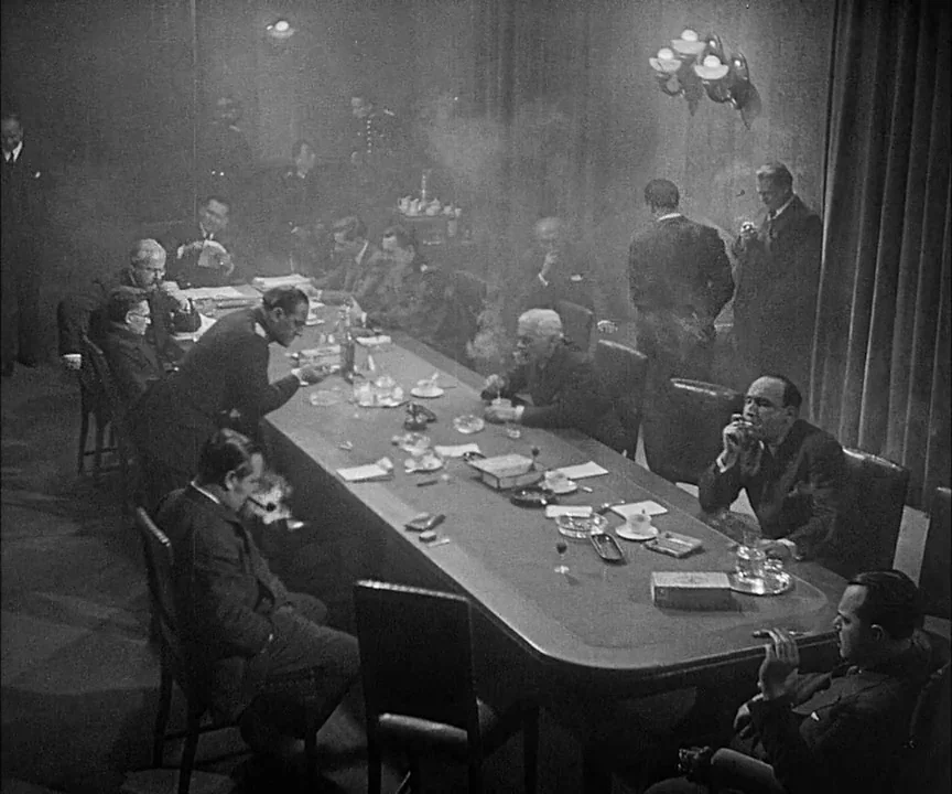 M (1931) movie still: wide — A smoke-filled 1940s conference room contains a large oval table with many seated men. The men sit around the table…