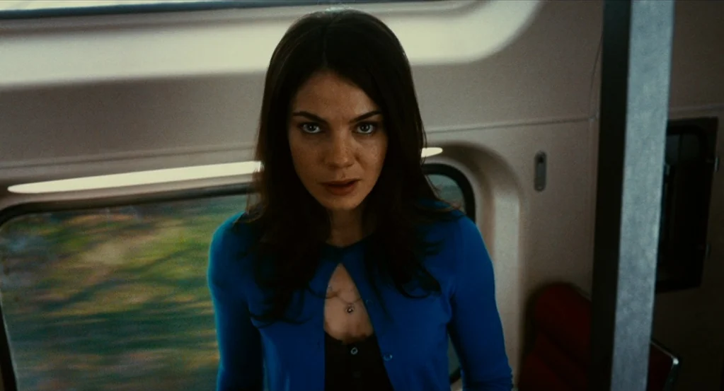 Source Code (2011) movie still: medium — A woman stands inside a passenger train car with windows blurred by motion. She looks ahead directly into…