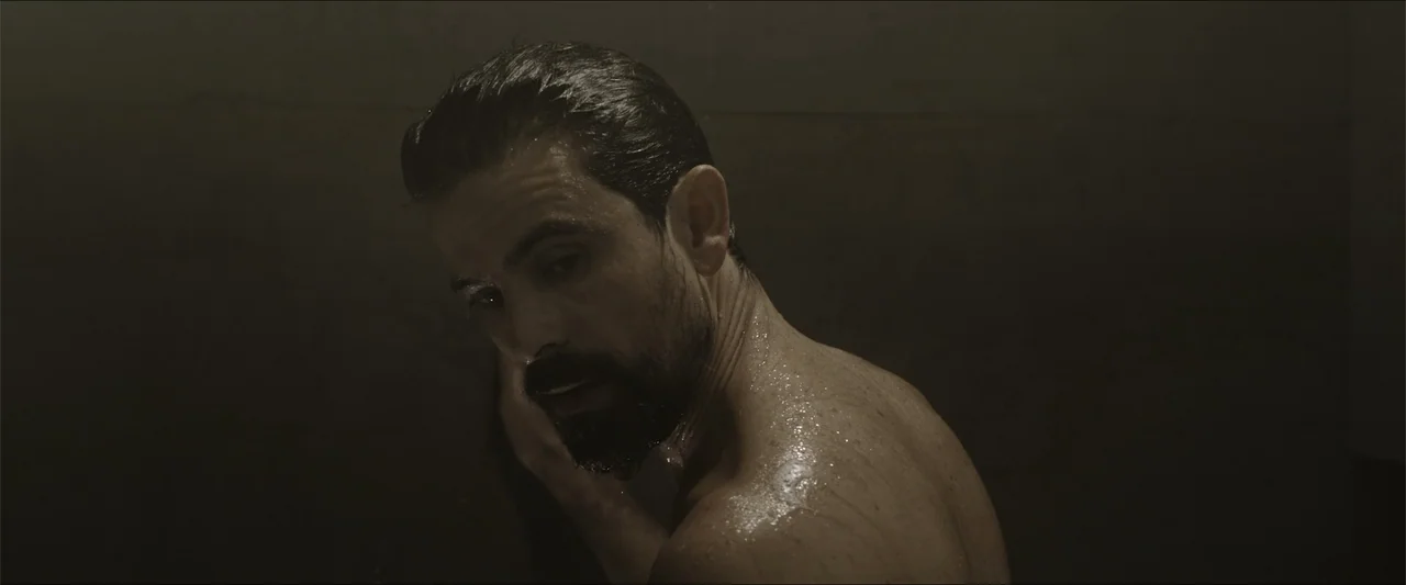 Los habitantes (2023) movie still: closeup — A dark, tiled interior space holds a person within a dimly lit shower area. A man with wet hair and…