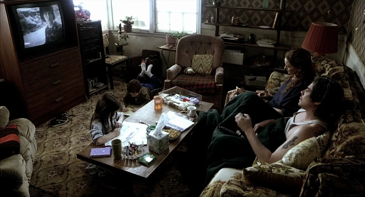 21 Grams (2003) movie still: wide — A cluttered suburban room features patterned wallpaper and a heavily used floral-patterned sofa. Two children…