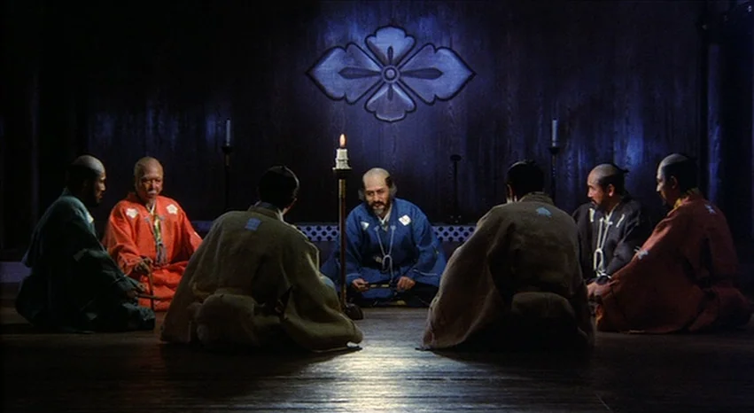 Kagemusha (1980) movie still: wide — A dark wooden room features a prominent crest mounted on the wall behind the group. Seven men in traditional…