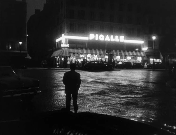 Bob le Flambeur (1956) movie still: wide — A wet Parisian intersection at night features a brightly lit neon Pigalle sign above storefronts. A lone…
