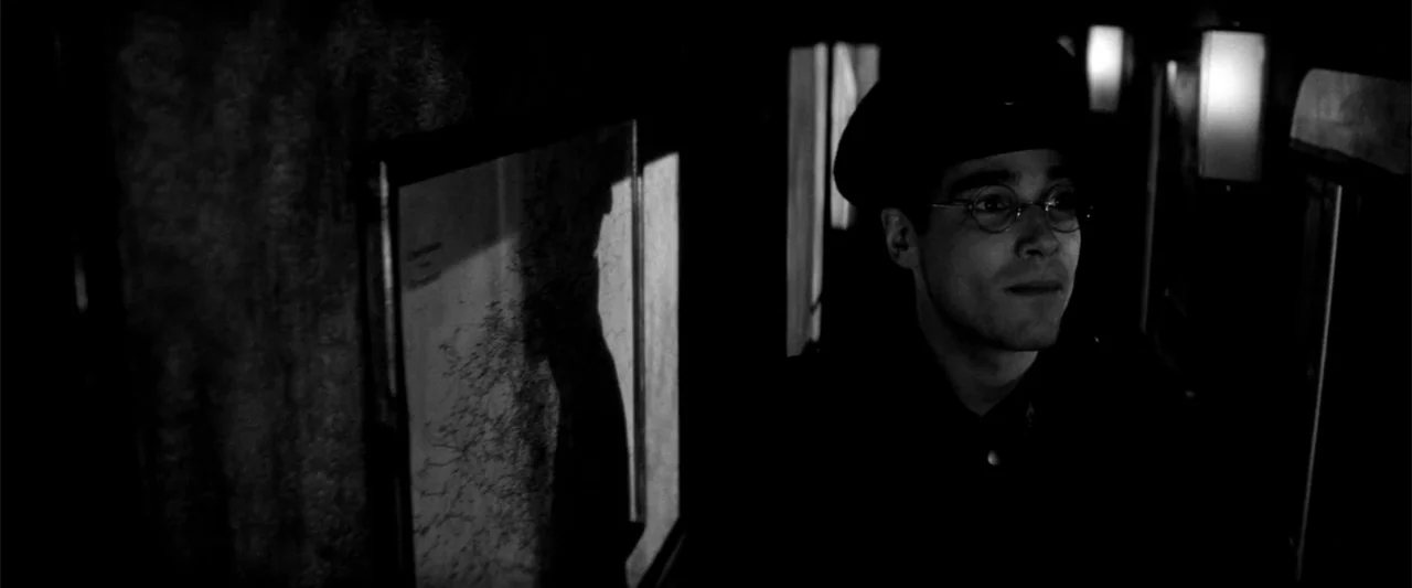 Europa (1991) movie still: medium — The interior of a dark, narrow train carriage with a wall-mounted framed map. A young man in a uniform and…
