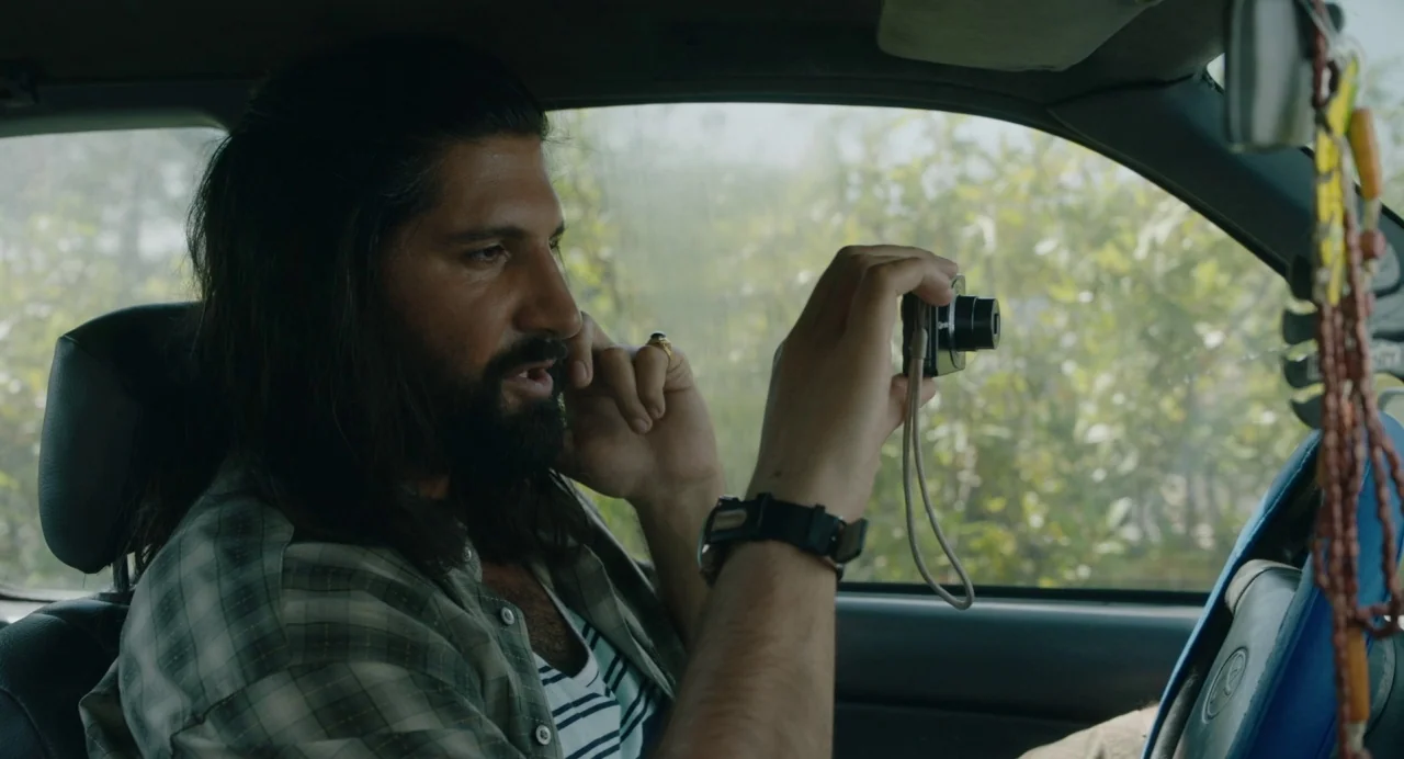 The Day Shall Come (2019) movie still: medium — A man sits inside a vehicle with trees visible through the window. The subject holds a small camera…