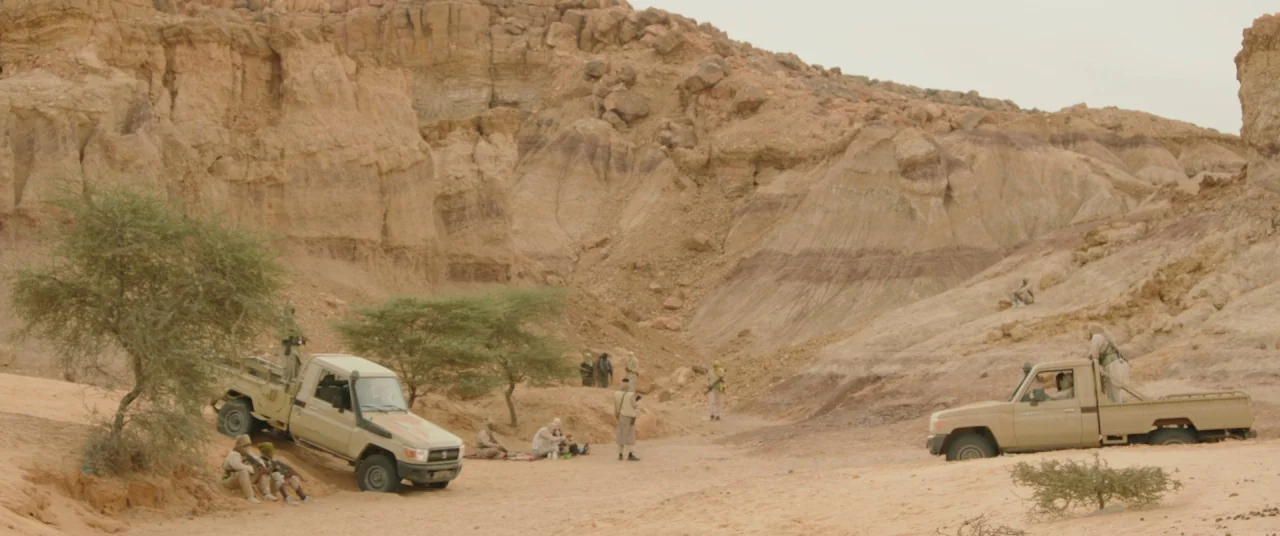 Timbuktu (2014) movie still: wide — An arid, rocky canyon landscape features high, eroded sandstone walls and sparse desert brush. A group of men in…