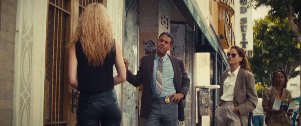 MaXXXine (2024) movie still: fullbody — An urban sidewalk outside a shop entrance featuring a brass door frame and signs. A man in a suit engages in…