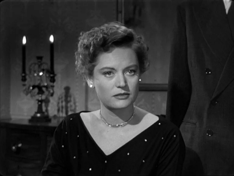 The Sleeping Tiger (1954) movie still: medium — A dimly lit room contains a decorative candelabra on a wooden surface. A woman in a black top sits…