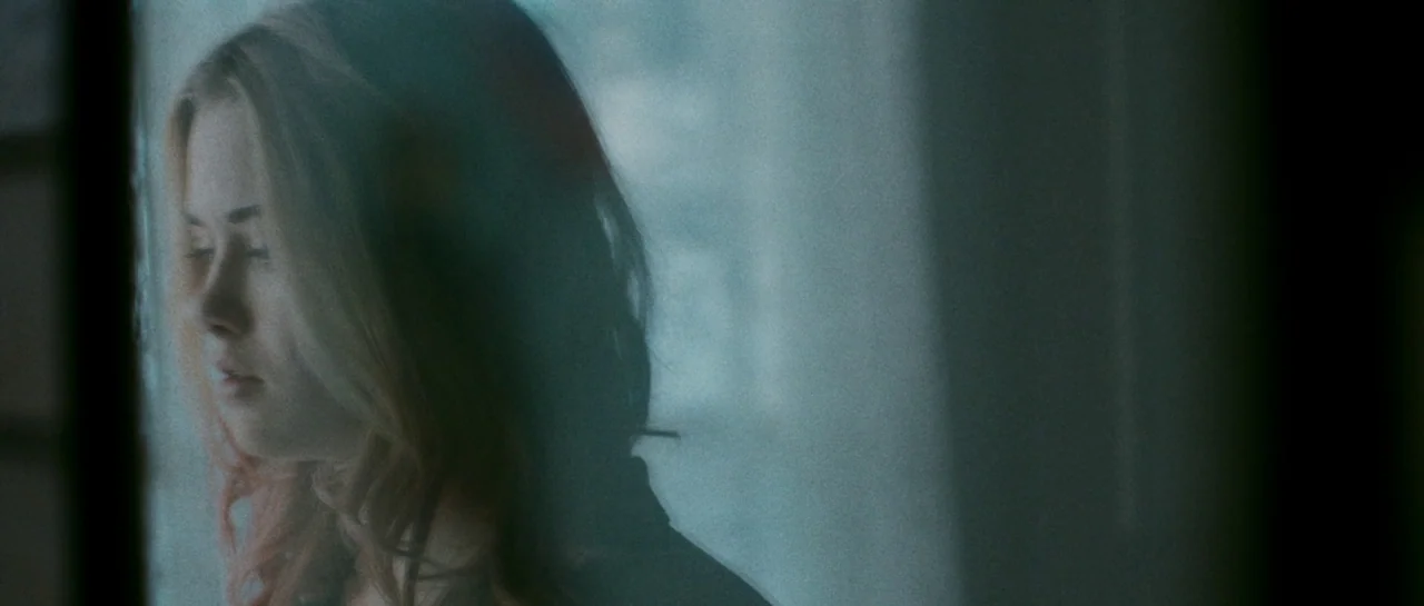 Starfish (2018) movie still: closeup — A close-up view of a young woman standing beside a window. Light reflects off the glass surface, partially…