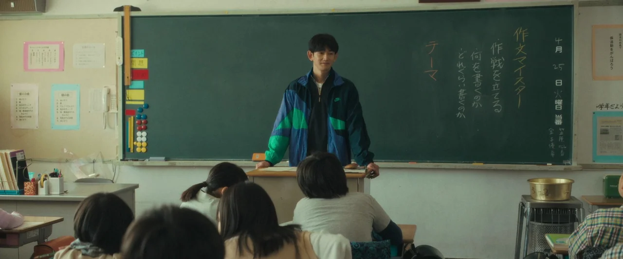 Monster (2023) movie still: medium — A Japanese classroom with a large green chalkboard covered in handwritten white and red text. A male teacher…