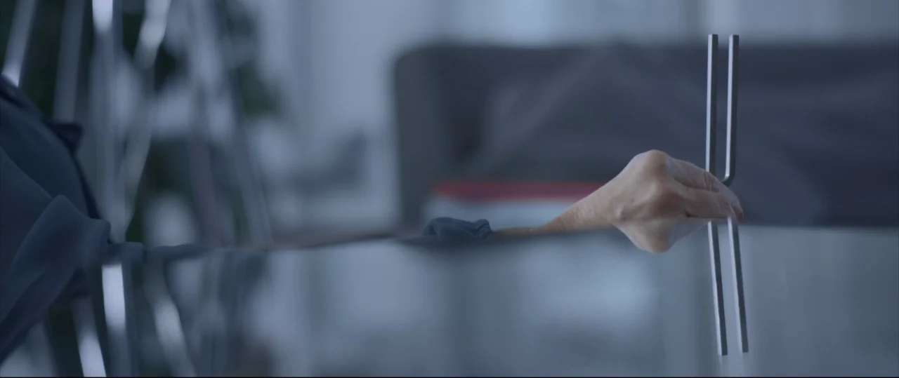 Sun Choke (2015) movie still: closeup — A quiet indoor setting features a glass tabletop and a blurred interior background. An elderly person's hand…