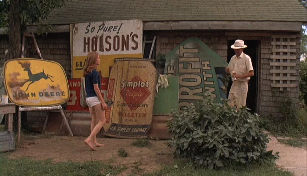 Badlands (1973) movie still: fullbody — A wooden shingled building exterior is cluttered with various vintage metal advertising signs. A barefoot…