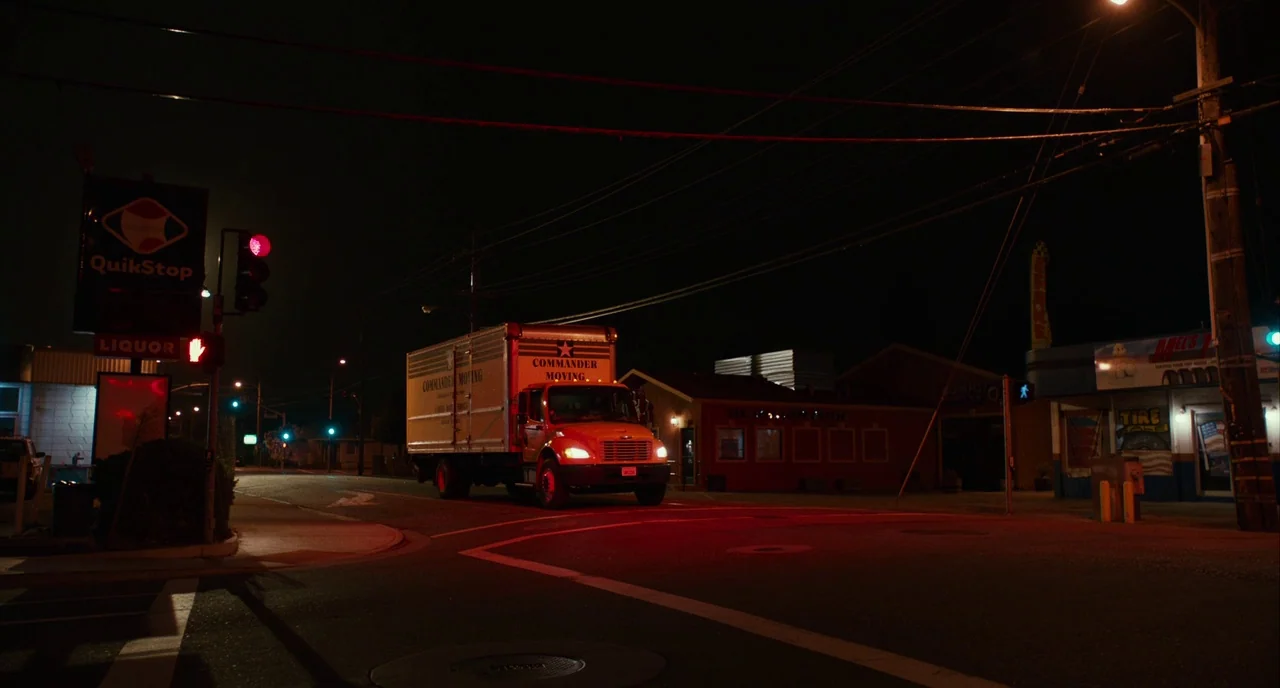 Blindspotting (2018) movie still: wide — A suburban street intersection at night features a moving truck turning under traffic lights. The…