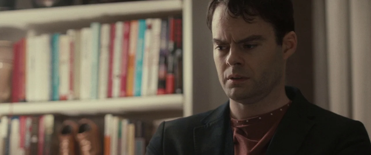 The Skeleton Twins (2014) movie still: medium — A man stands before a white bookshelf filled with numerous books arranged horizontally. He faces…