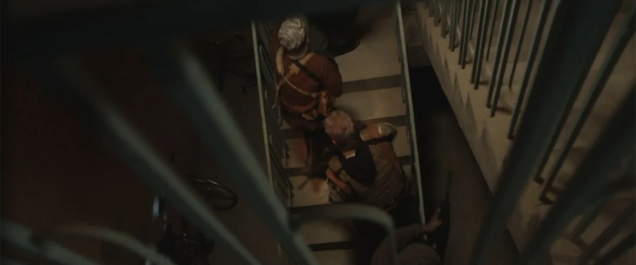 Presagios (2024) movie still: medium — A dimly lit concrete stairwell with industrial railings and shadows cast by low lighting. Two men in tactical…