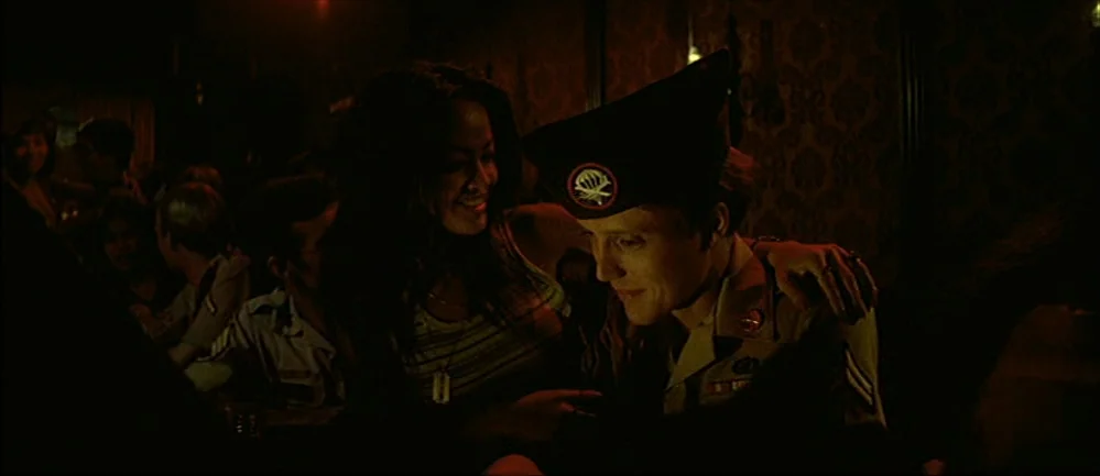 The Deer Hunter (1978) movie still: medium — A dark, wood-paneled bar with ornate wallpaper and crowded seating in the background. A woman in a…