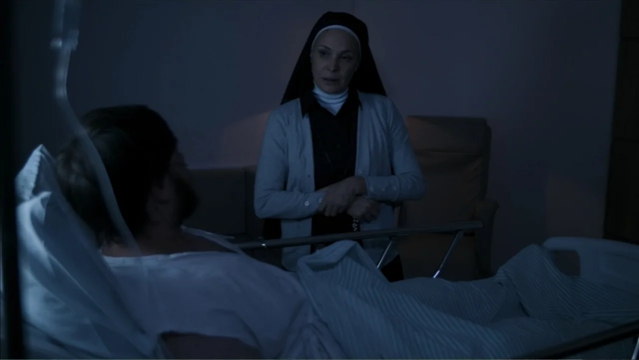 The Grotto (2022) movie still: medium — A dark hospital room at night features a man in a bed and a nun. The nun stands and looks towards the man…