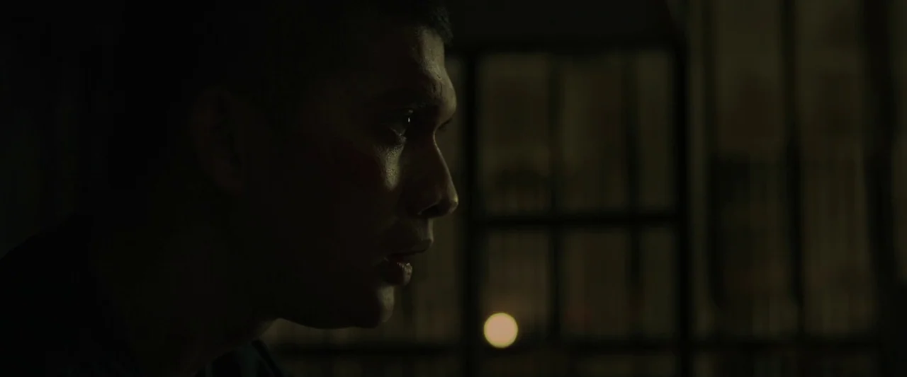 The Raid 2 (2014) movie still: closeup — A dark, dimly lit prison cell features metal bars visible in the background shadow. A man stands in the…