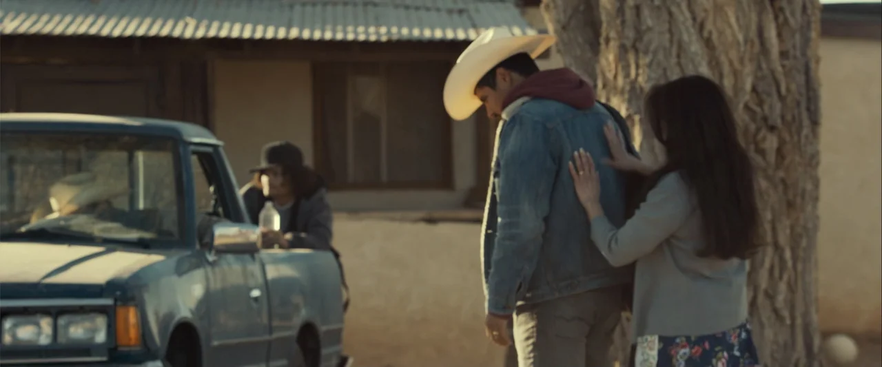 Frontera (2014) movie still: medium — An exterior dusty lot next to a simple building and a parked pickup truck. A woman stands behind a man in a…