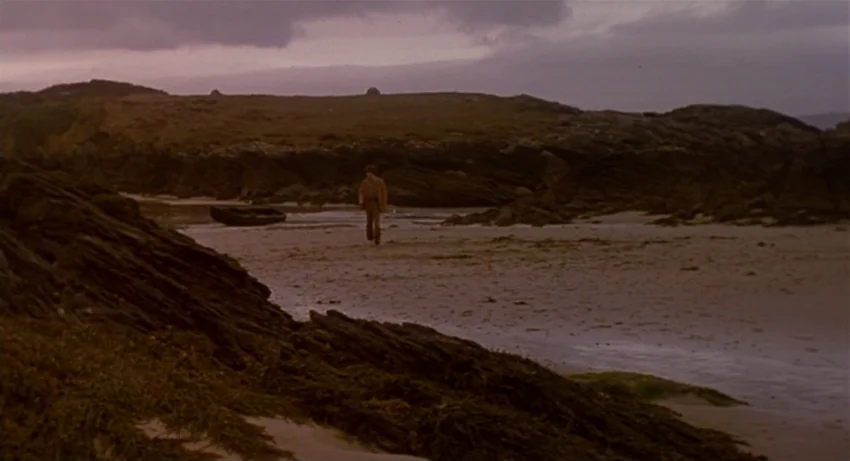 The Secret Of Roan Inish (1994) movie still: wide — A dark rocky shoreline with wet sand stretches beneath a heavy overcast sky. A lone person walks…