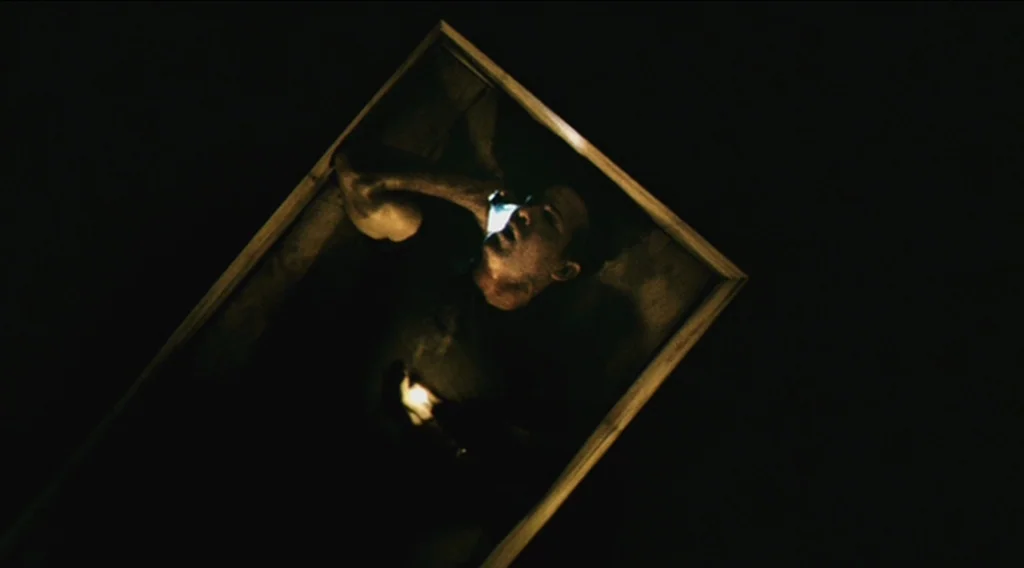 Buried (2010) movie still: closeup — A man lies in a dark, enclosed space framed by a light-colored wooden structure. He holds a bright mobile phone…
