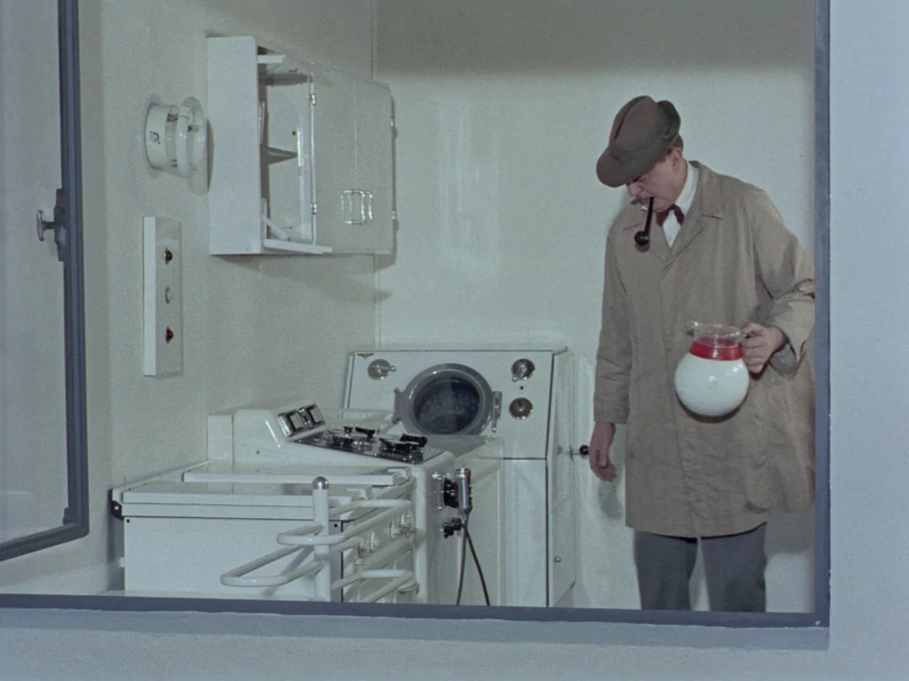Mon Oncle (1958) movie still: medium — A stark white room contains a retro stove and mounted cabinets on the wall. A man in a trench coat and flat…