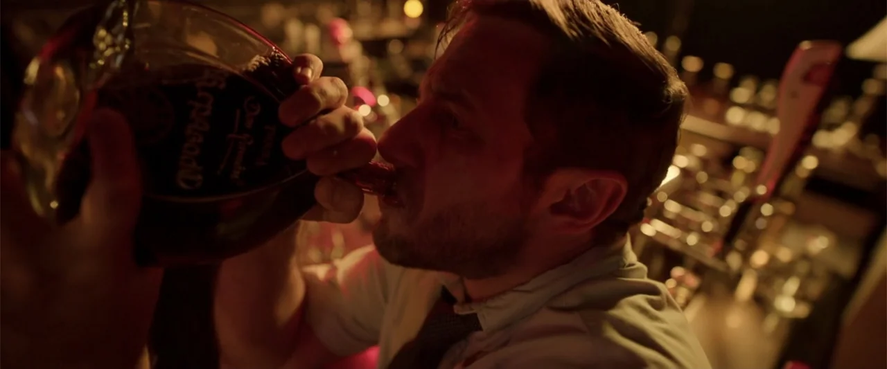 Tequila Re-Pasado (2023) movie still: closeup — A dimly lit bar interior contains a man holding a bottle. He tilts his head back to drink from the…