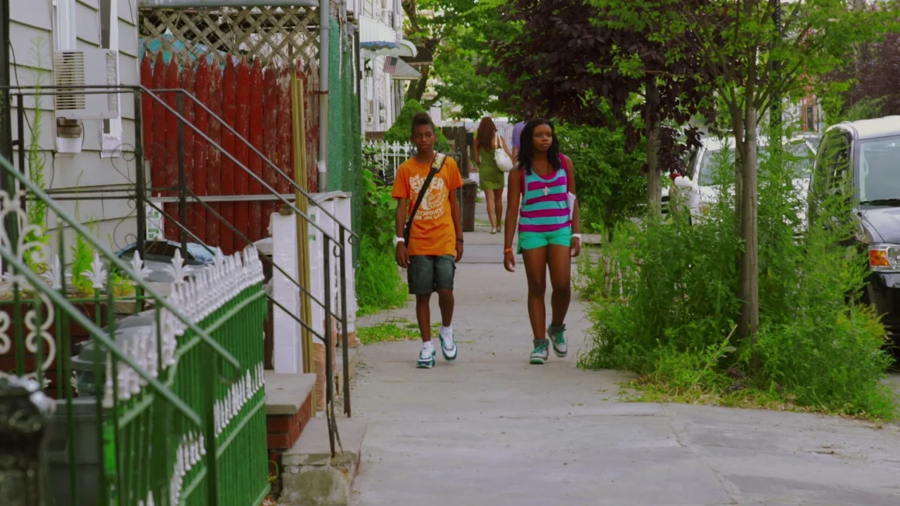 Red Hook Summer (2012) movie still: fullbody — A residential sidewalk runs alongside a house with a white fence and overgrown greenery. Two…
