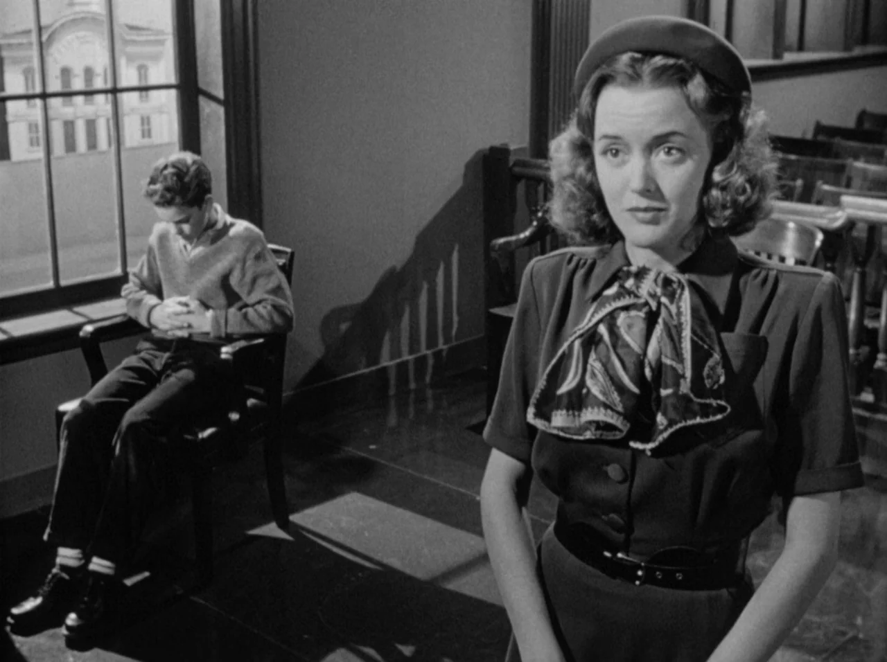 Gun Crazy (1950) movie still: medium — A quiet indoor space features large windows overlooking a building across the street. A woman stands in the…