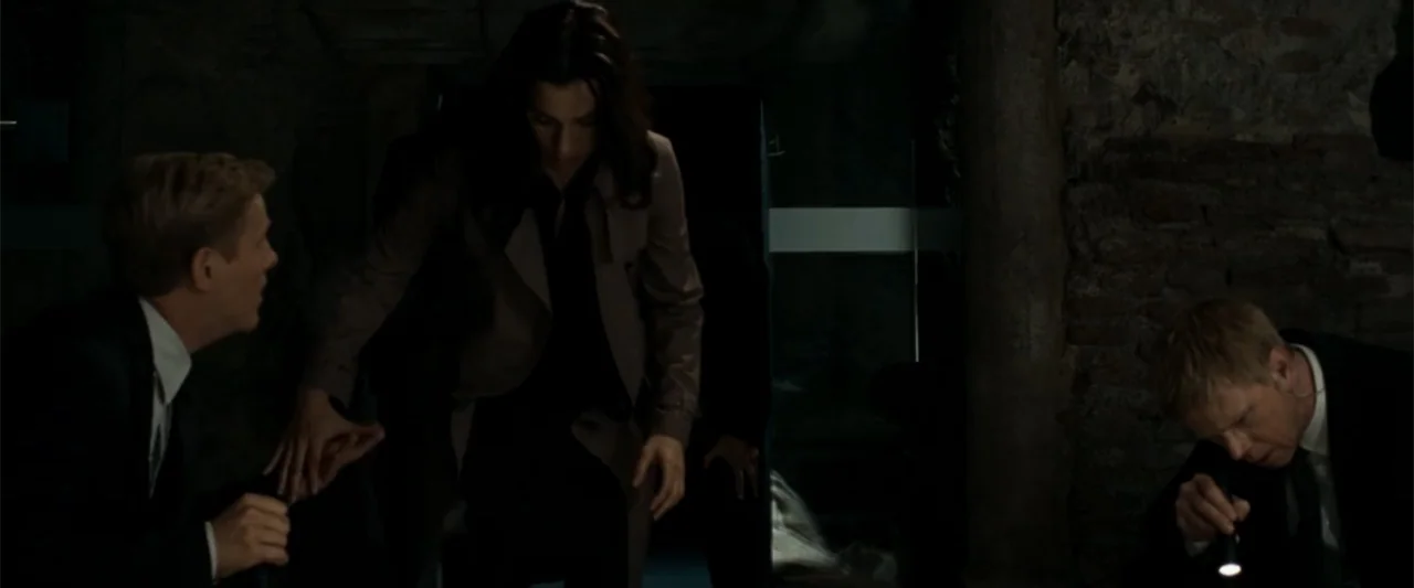 Angels & Demons (2009) movie still: medium — Three individuals explore a dark subterranean stone chamber with exposed brick walls. The group moves…