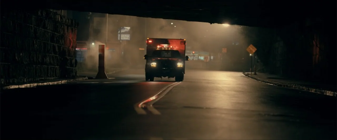 Madame Web (2024) movie still: wide — An asphalt road runs through a dark stone underpass at night with thick haze. An ambulance with flashing…