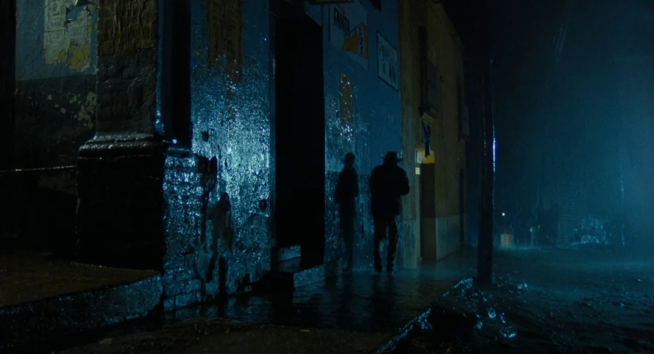 Canoa: A Shameful Memory (1976) movie still: wide — A dark urban exterior with blue-tinted walls and wet pavement at night. Two men move along the…