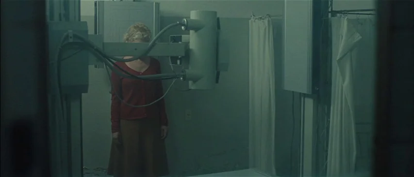 The Headless Woman (2008) movie still: medium — A medical imaging room contains an x-ray machine and white fabric privacy curtains. The woman stands…