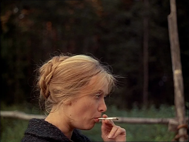 Mirror (Зеркало, Zerkalo) (1975) movie still: closeup — A woman stands outdoors in front of a simple wooden fence and dark trees. She holds a lit…