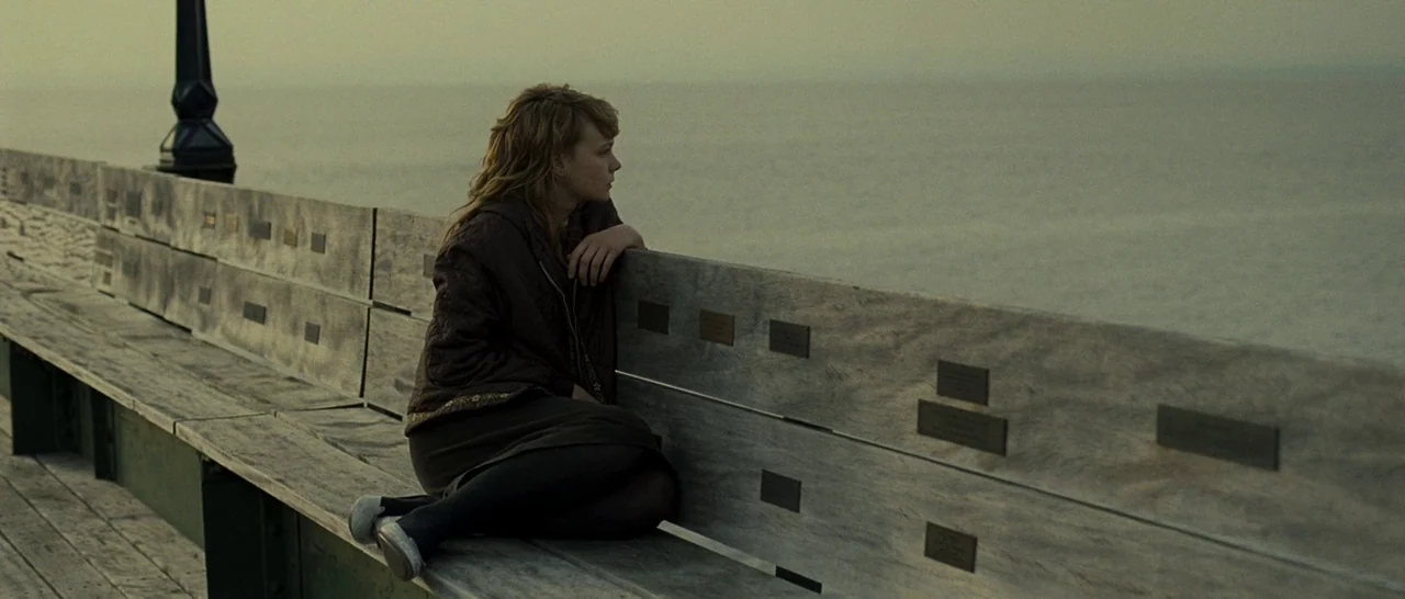 Never Let Me Go (2010) movie still: medium — A long wooden bench on a pier overlooks a calm, gray sea under overcast skies. A young woman sits on…