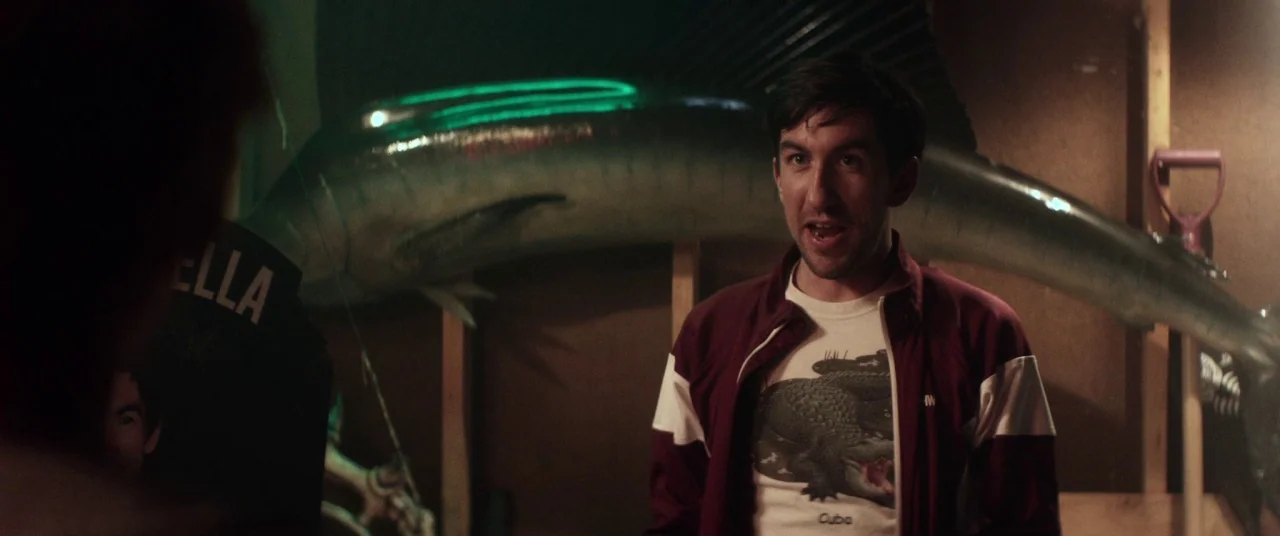 Room For Rent (2017) movie still: medium — A dimly lit garage interior features wood panel walls and a taxidermied fish mount. A man stands in the…